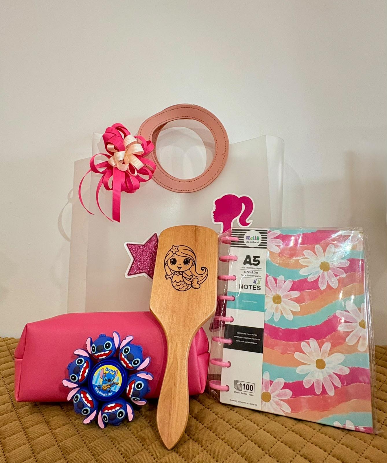 A Barbie-themed stationery gift set arranged on a quilted tan surface against a plain off-white wall. The set includes a clear gift bag with pink handles and a pink bow, a pink notebook, a wooden spatula engraved with a Barbie mermaid, a pink hairbrush, and a pink floral spiral notebook.